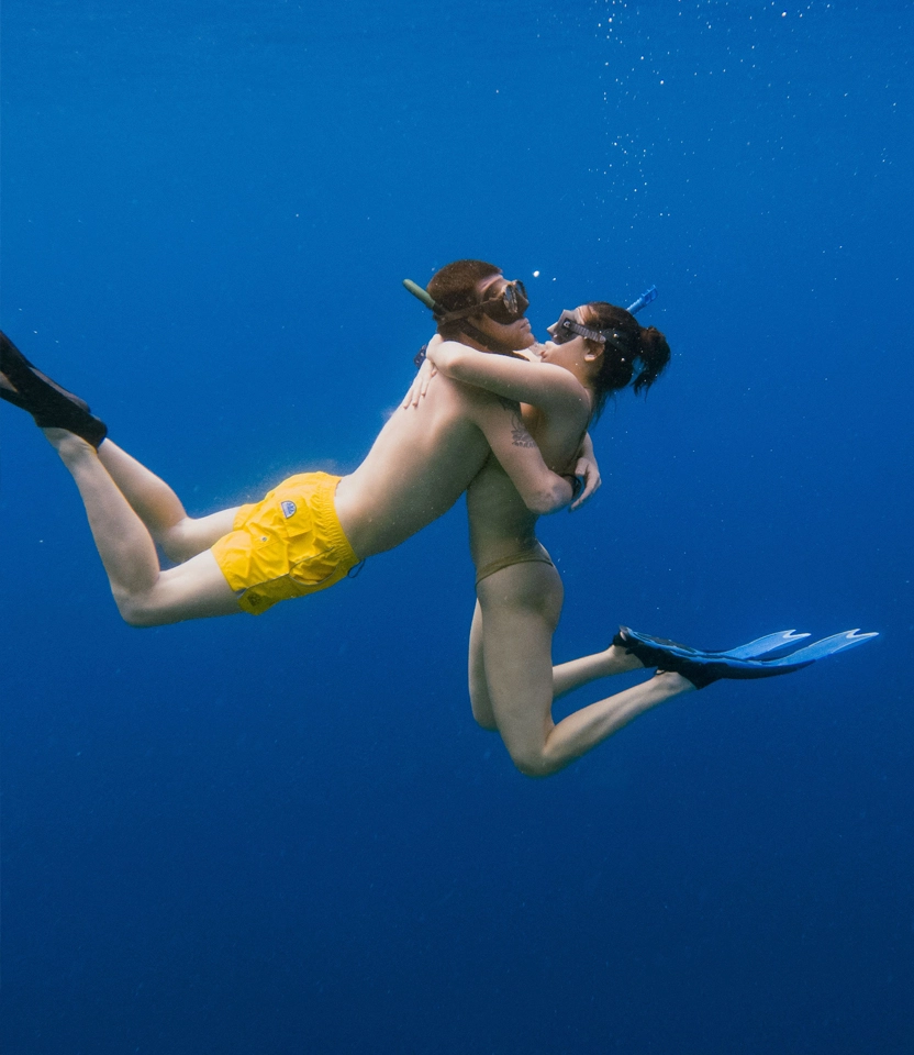 Um casal apaixonado se abraçando e se beijando embaixo d'água enquanto pratica snorkeling no mar azul da Ilha do Xavier, em Florianópolis. Eles estão usando máscaras e nadadeiras.